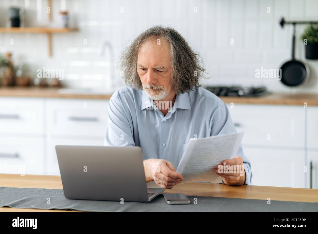 Ein enttäuschter, verärgerter, grauhaariger Kaukasier mittleren Alters sitzt in der Küche an einem Tisch mit Laptop, hält Dokumente in seinen Händen, erledigt Papierkram, gleicht Daten ab, analysiert die Ergebnisse der Arbeit Stockfoto