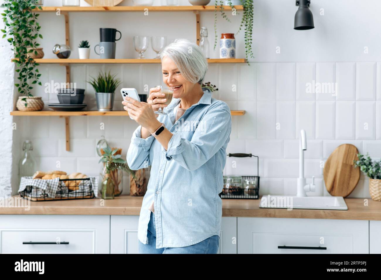 Fröhliche, weiße, stilvolle, grauhaarige Frau mittleren Alters, steht zu Hause in der Küche, hält Smartphone und ein Glas Kaffee, schickt Nachrichten in sozialen Netzwerken, liest Nachrichten, nimmt ein Selfie, lächelt Stockfoto