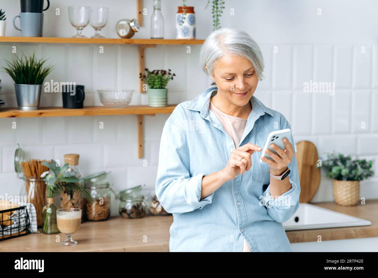 Erstaunliche moderne, weiße, reife, grauhaarige Frau, steht zu Hause in der Küche, hält ihr Smartphone in der Hand, benutzt es, um in sozialen Netzwerken zu kommunizieren, Nachrichten zu lesen, im Internet zu surfen, zu lächeln Stockfoto
