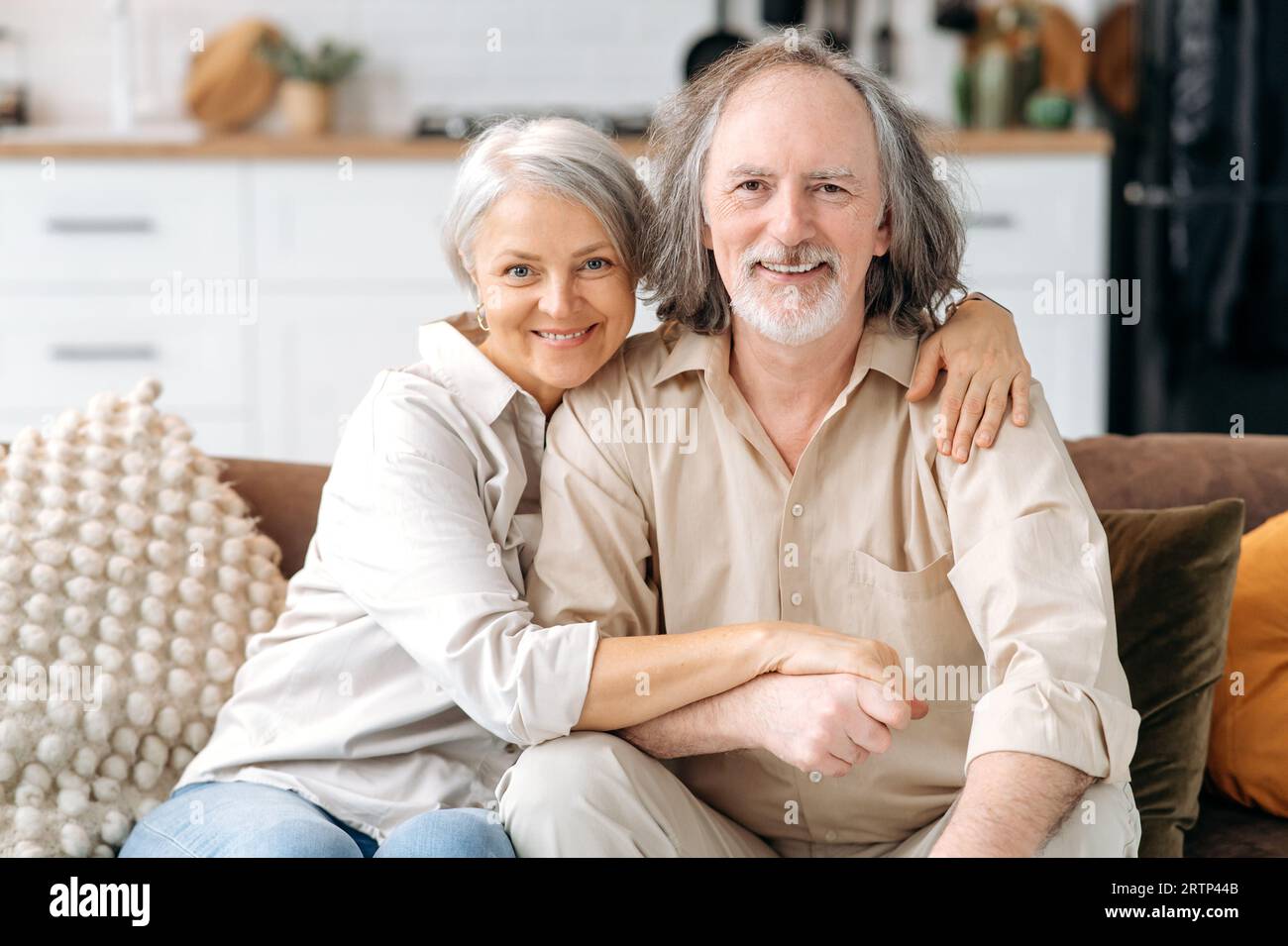 Weißes Ehepaar mittleren Alters, grauhaarige Ehepartner verbringen Zeit zusammen im Wohnzimmer zu Hause, Frau und Ehemann sitzen auf dem Sofa, umarmen sich, schauen in die Kamera, lächeln. Porträt eines glücklichen Rentners Stockfoto
