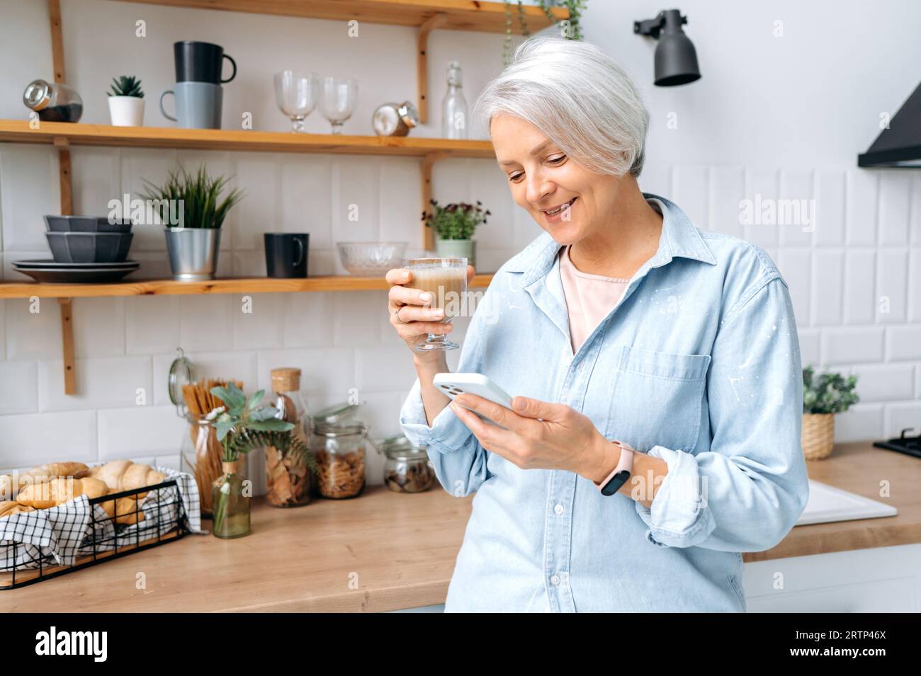 Zufriedene weiße Frau mittleren Alters mit grauhaarigen Haaren, steht zu Hause in der Küche, hält ihr Smartphone und ein Glas Kaffee, chattet in sozialen Netzwerken, liest Nachrichten, macht eine Online-Bestellung, lächelt Stockfoto