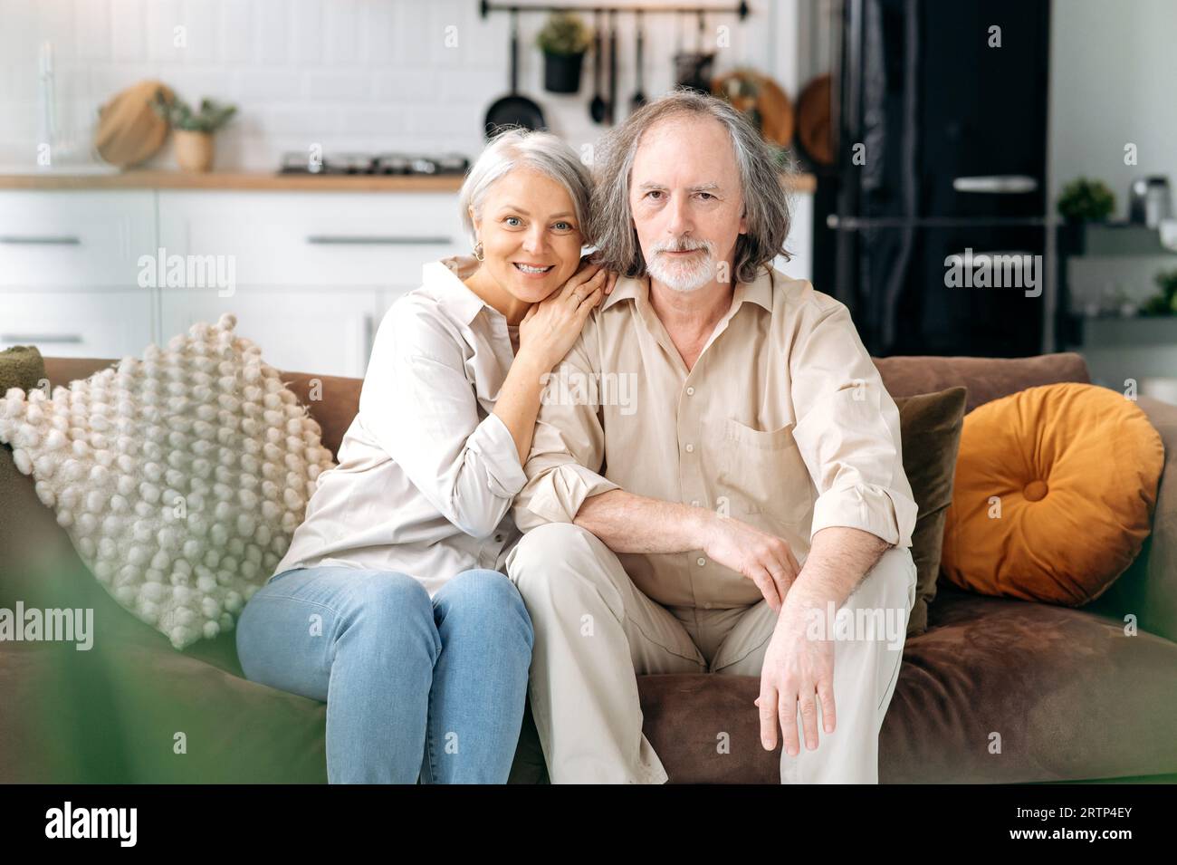 Glücklich zusammen. Moderne Rentner. Weißes Ehepaar mittleren Alters, grauhaarige Ehepartner verbringen Zeit zusammen im Wohnzimmer, Frau und Ehemann sitzen auf dem Sofa, umarmen sich, schauen in die Kamera, lächeln Stockfoto