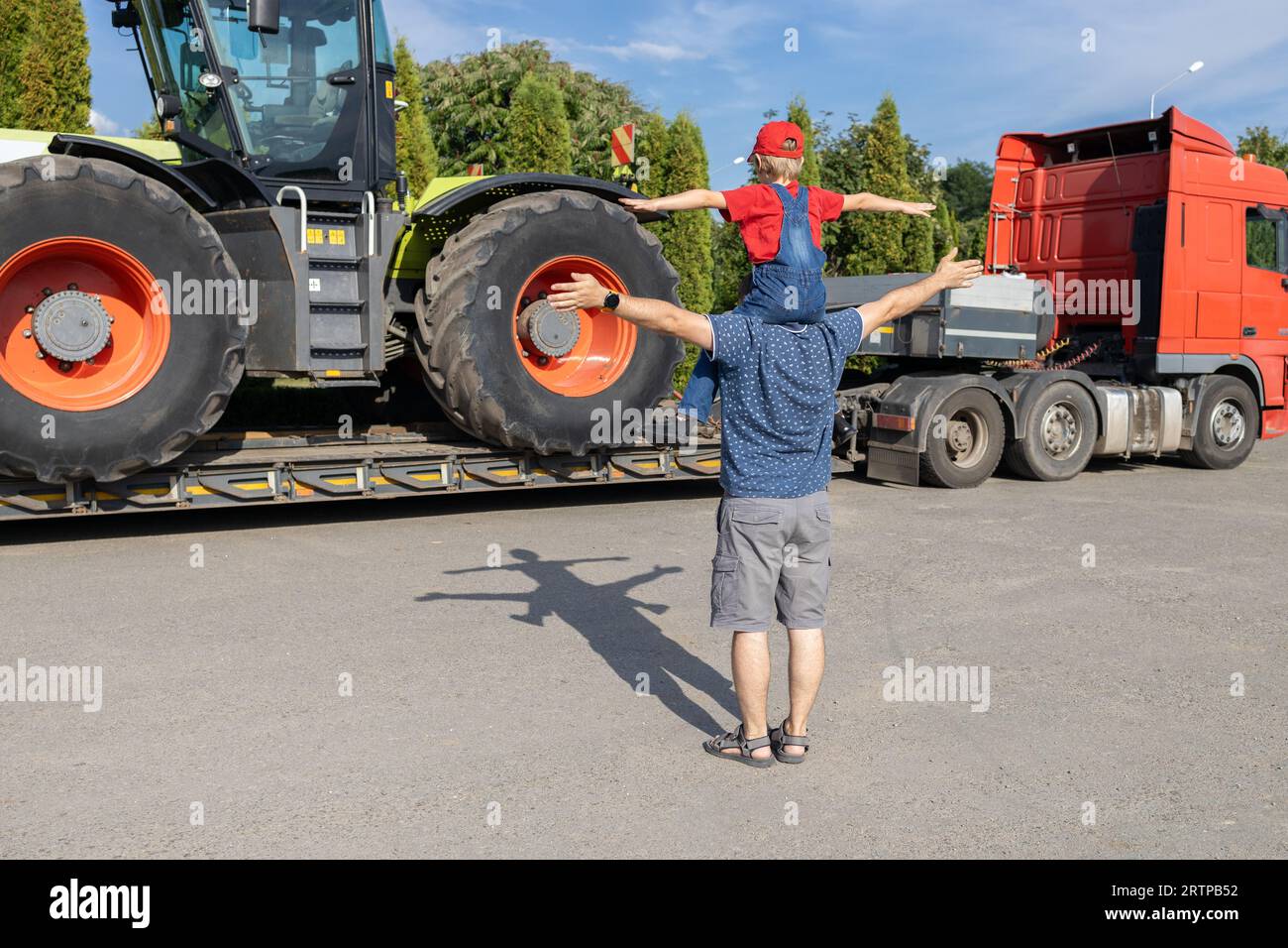 Mann und Junge stehen vor einem großen Traktor, der auf einem Lkw-Anhänger steht, Rückansicht. Die Leidenschaft des Sohnes für Autos, Papa und Kind schauen sich Fahrzeuge in der Parkette an Stockfoto
