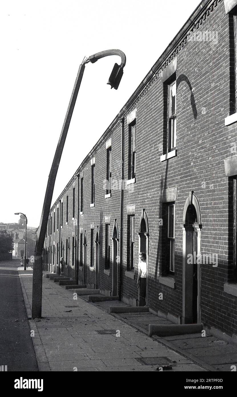 1960er Jahre, historisch, draußen in der Straße von viktorianischen Terrassenhäusern, eine Dame in ihrem Eingang, die auf einen Biegeleuchte Pfosten gegenüber ihrem Haus, Ripponden Rd, Oldham, England, Großbritannien, blickt. Stockfoto