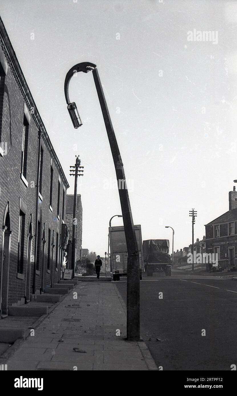 1960er Jahre, historisch, ein geknickter Laternenpfahl in der Straße mit kleinen viktorianischen Reihenhäusern, Ripponden Rd, Oldham, England, Großbritannien. Gegenüber einem Wilson's Pub, dem Bulls Head. Stockfoto