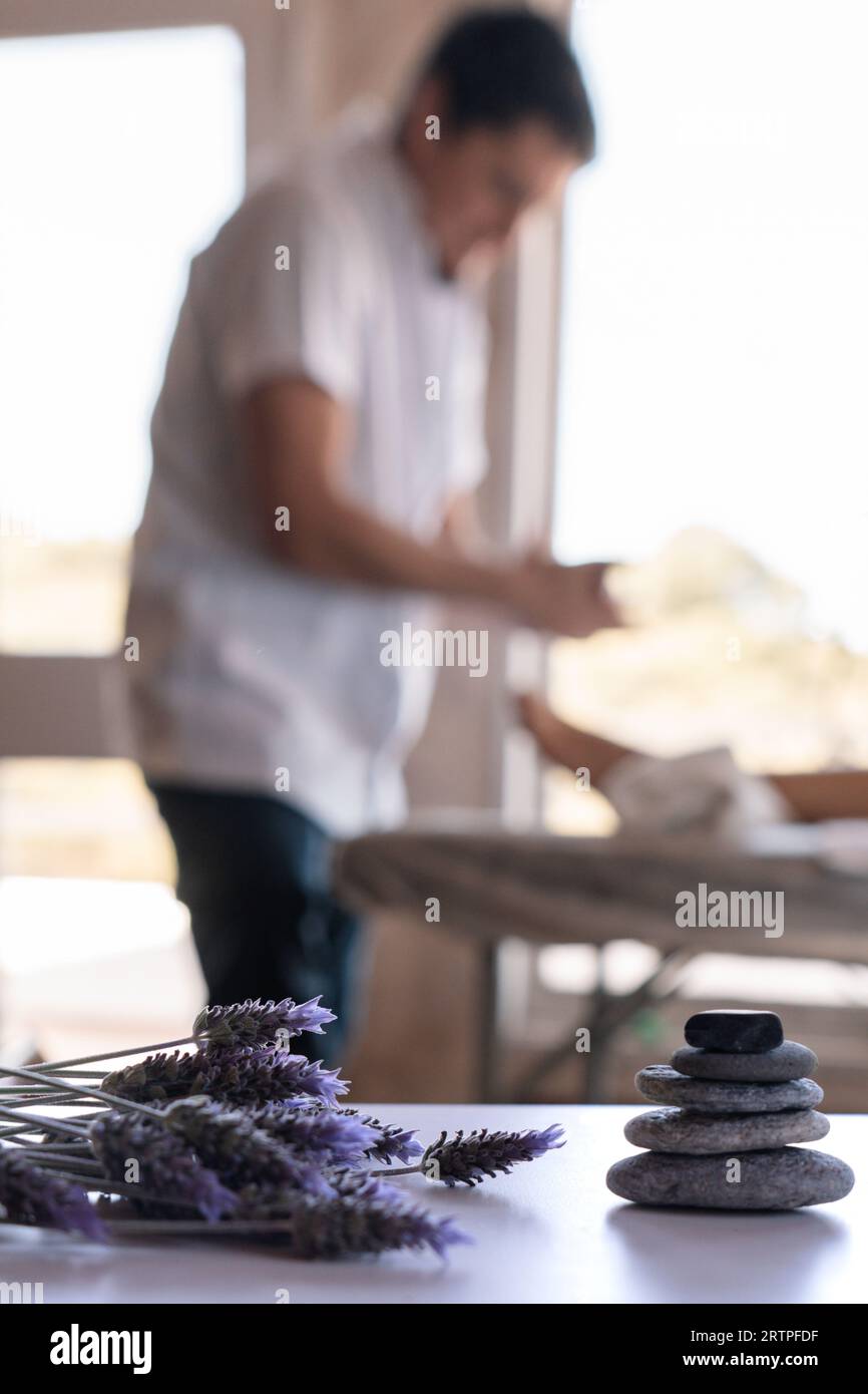 Verschwommener Hintergrund eines professionellen männlichen Masseurs und seines Patienten in einem Spa, im Vordergrund einige Lavendelblüten und ein Stein, der auf jeden OT gestapelt ist Stockfoto