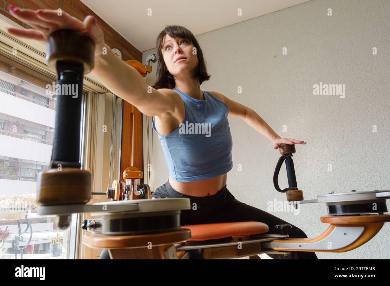 Glückliche Latina Frau mit kurzen schwarzen Haaren, in Sportkleidung, sitzend mit einem Arm hinter und einer vorne Training zu Hause mit hölzerner Übung und Stretch Stockfoto