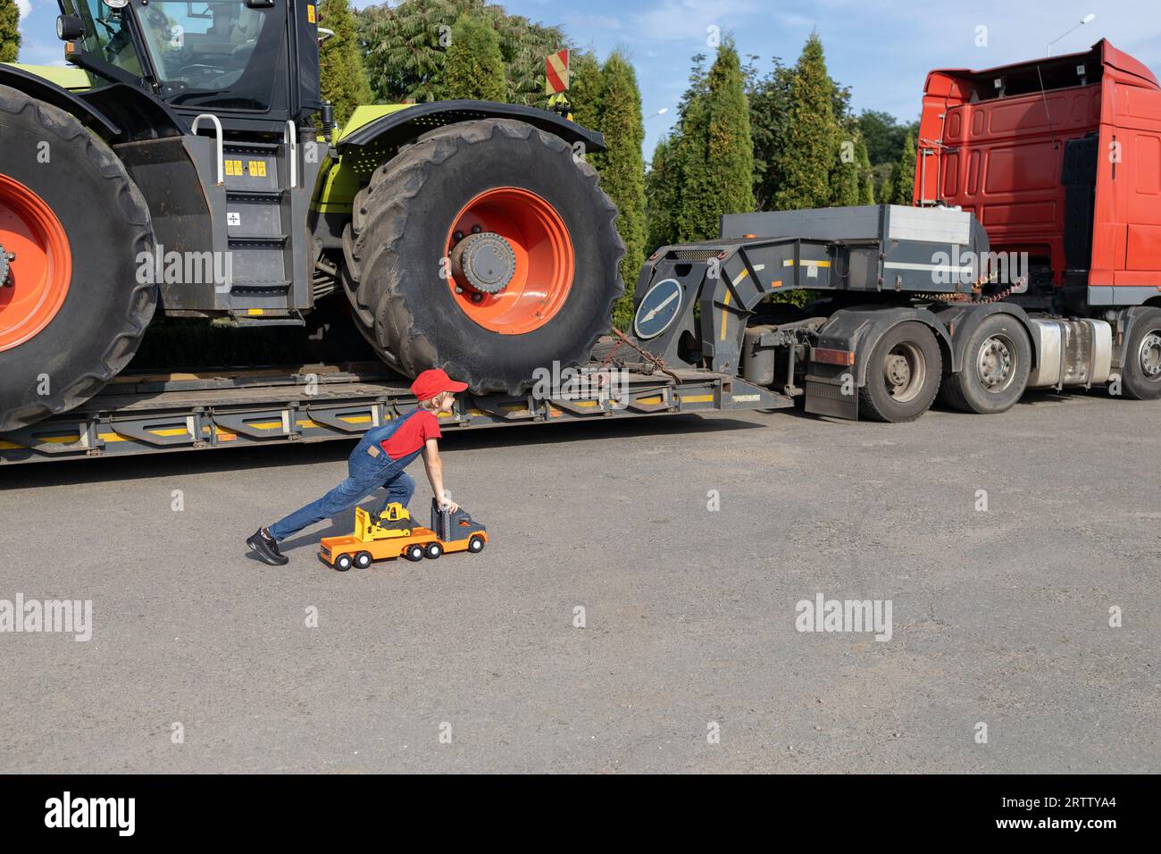 Der Junge spielt mit dem Kipplaster. Hinter dem Kind befindet sich ein großer Lkw mit beladenem Traktor. Träume, ein Fahrer oder Mechaniker zu werden, wie Dad. Kommerziell V Stockfoto