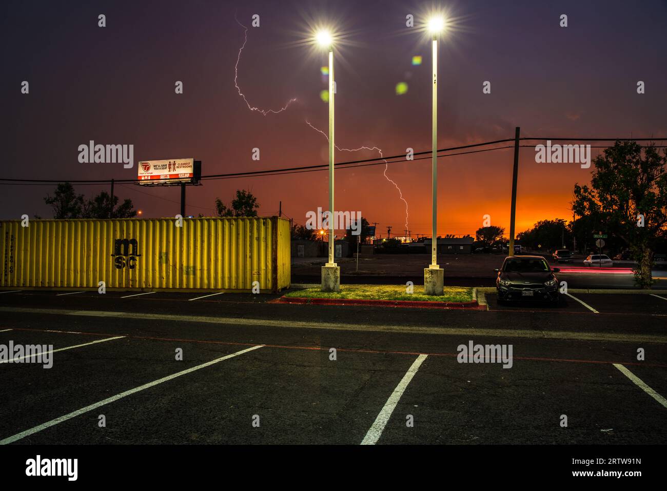 Blitze in der magischen Stunde hinter einem zufälligen Parkplatz mit einem leuchtend gelben Schiffscontainer, während der Tag in Amarillo, Texas, zur Nacht wird. Stockfoto