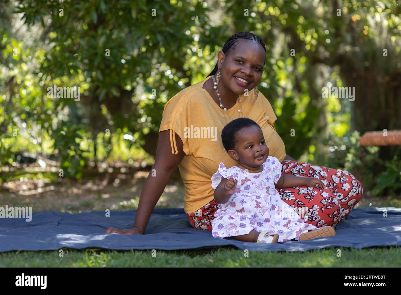 Porträt einer jungen schwarzen Frau, die auf einer Picknickdecke unter Bäumen mit ihrer kleinen Tochter sitzt Stockfoto