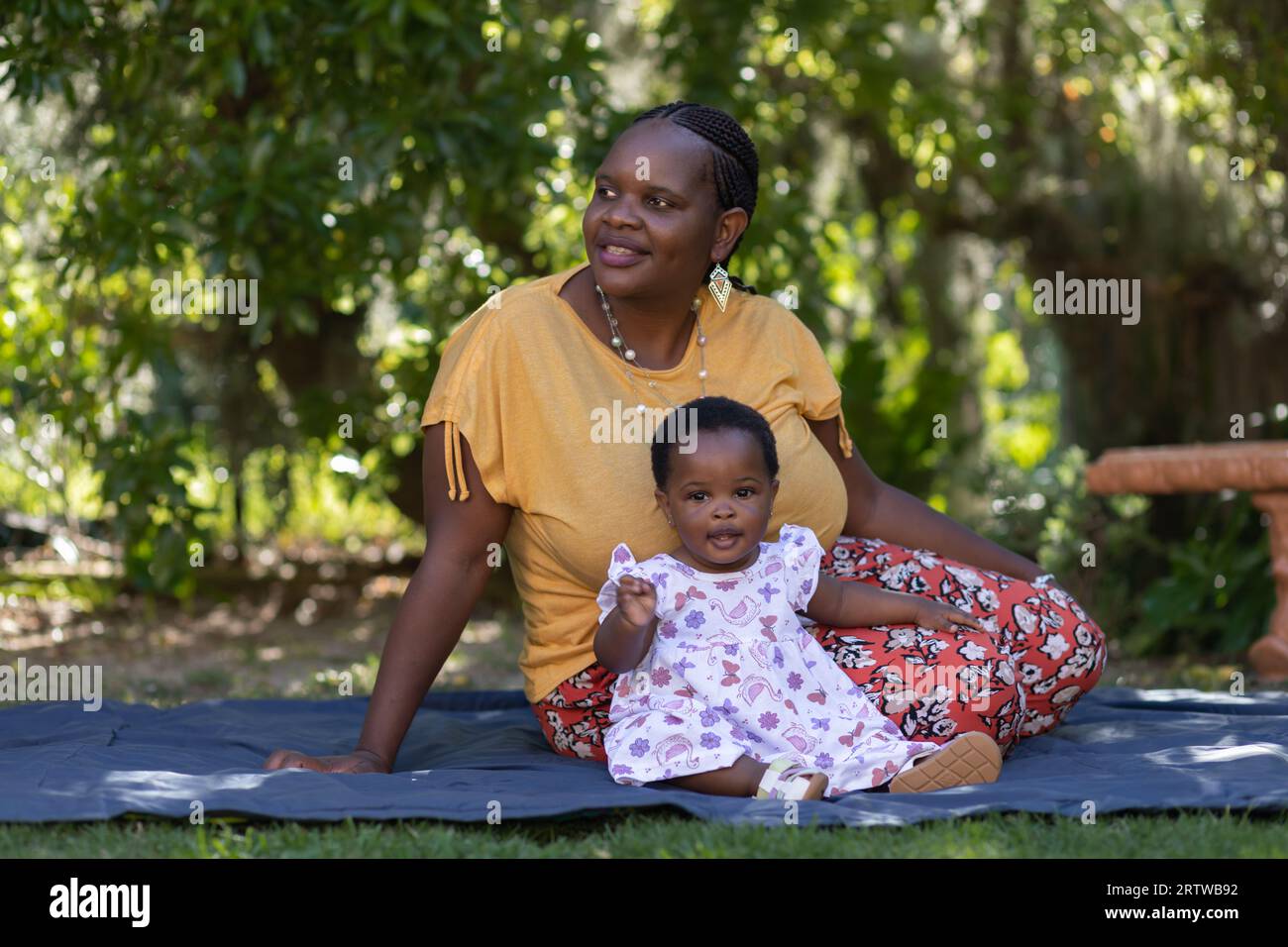 Porträt einer jungen schwarzen Frau, die auf einer Picknickdecke unter Bäumen mit ihrer kleinen Tochter sitzt Stockfoto