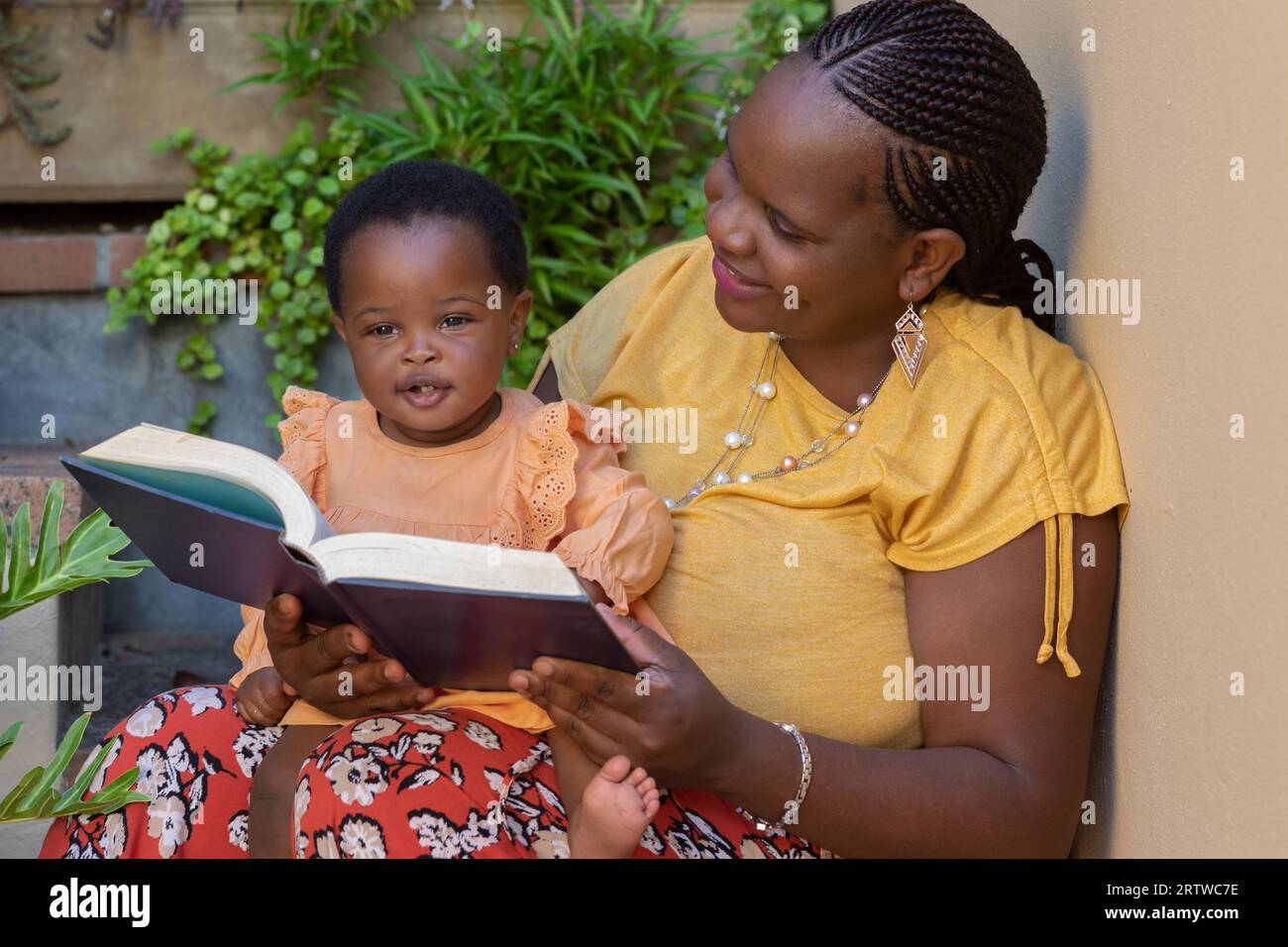 Porträt einer jungen schwarzen Mutter, die ihrer kleinen Tochter vorliest, die auf ihrem Schoß sitzt Stockfoto