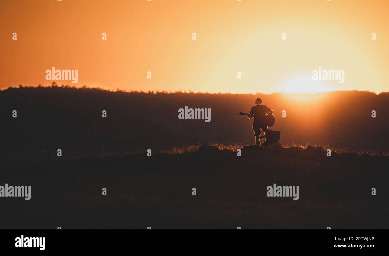 Silhouette eines Mannes, der bei Sonnenuntergang auf der Wiese Gitarre spielt. Hügel im Hintergrund. Orangefarbener Himmel. Abstrakte Ansicht. Stockfoto