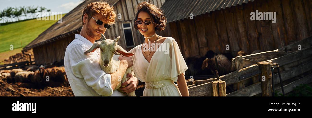 Lächelndes multiethnisches Paar im Hochzeitskleid und Sonnenbrille kuscheln Babyziege auf dem Land, Banner Stockfoto