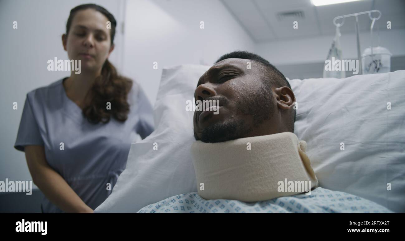 Junge Krankenschwester bringt verletzten Mann mittleren Alters auf Krankenhausstation. Der afroamerikanische Patient liegt auf einer trage in der Nackenstütze und spricht mit dem Arzt über die Gesundheit. Medizinisches Personal am Arbeitsplatz im medizinischen Zentrum. Stockfoto