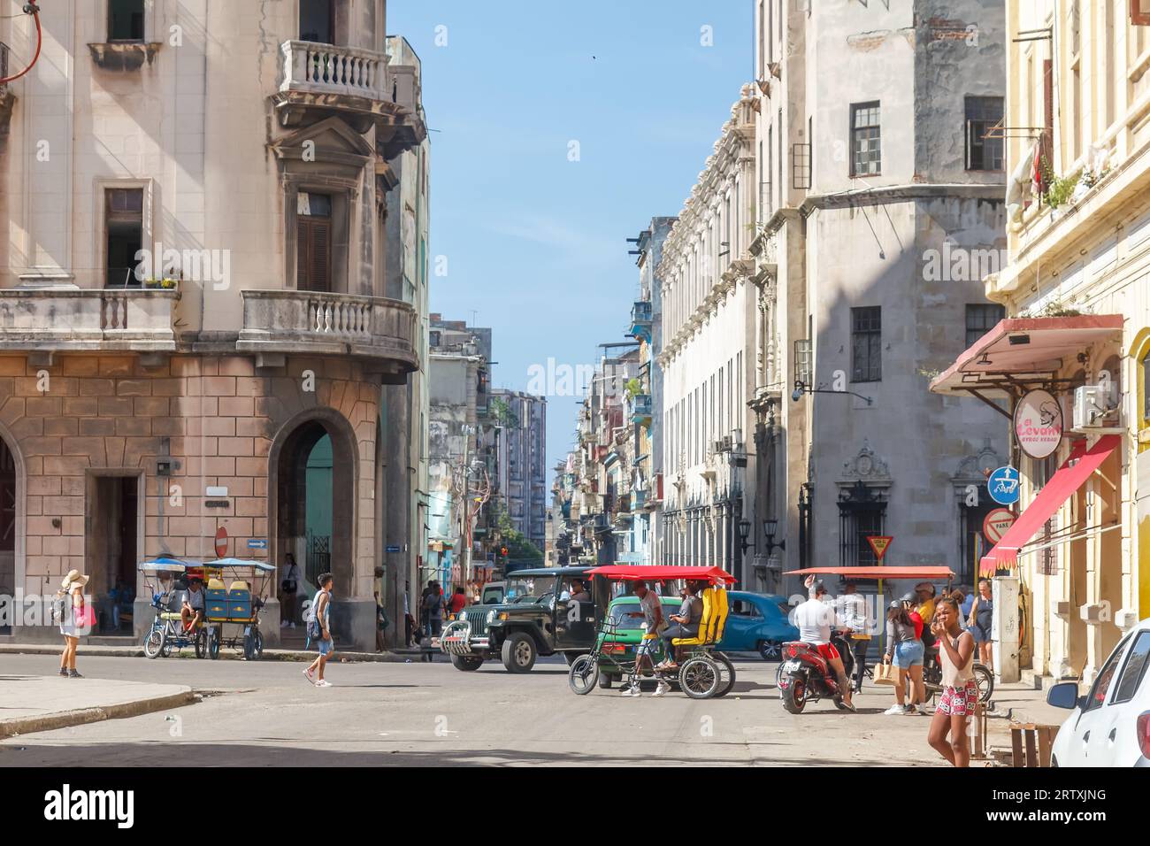 Havanna, Kuba, 2023, Bicitaxis und Kraftfahrzeuge fahren in einer Stadtstraße, wo verwitterte alte Gebäude an der Kreuzung zu sehen sind. Stockfoto