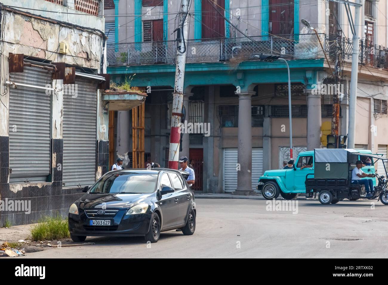 Havanna, Kuba, 2023: Ein modernes Auto steht im Kontrast zu alten Fahrzeugen in einer Stadtstraße – die traditionellen heruntergekommenen und verwitterten Gebäude stehen im Hintergrund. Stockfoto