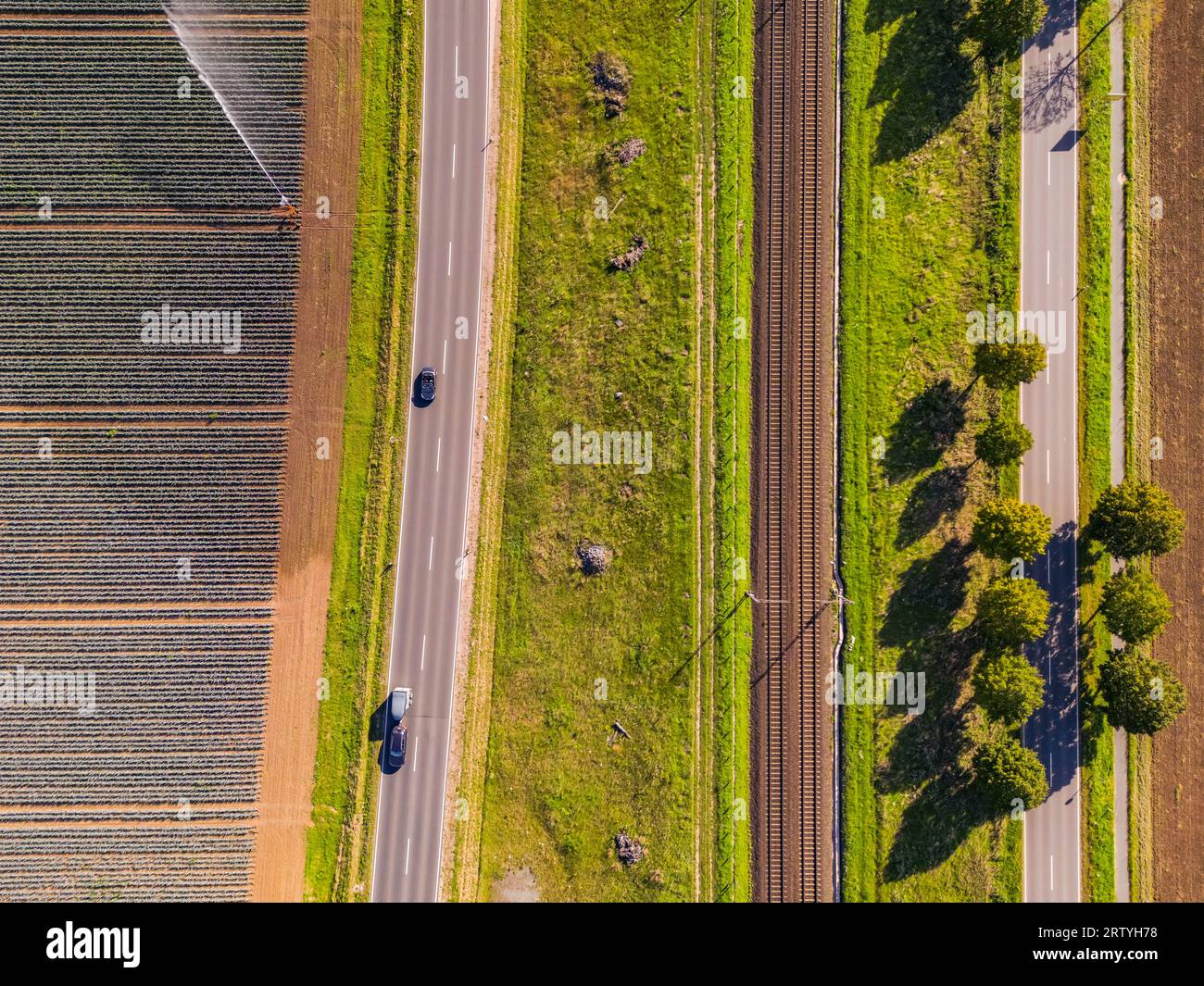 Eine Eisenbahnstrecke mit zwei Gleisen neben zwei Straßen und Feldern neben landwirtschaftlichen Feldern Stockfoto