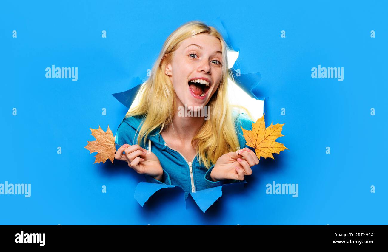 Glückliches blondes Mädchen mit herbstlichen Blättern, die durch ein Papierloch schauen. Herbstkollektion. Lächelnde Frau im warmen Pullover mit Ahornblatt. Lässig Stockfoto