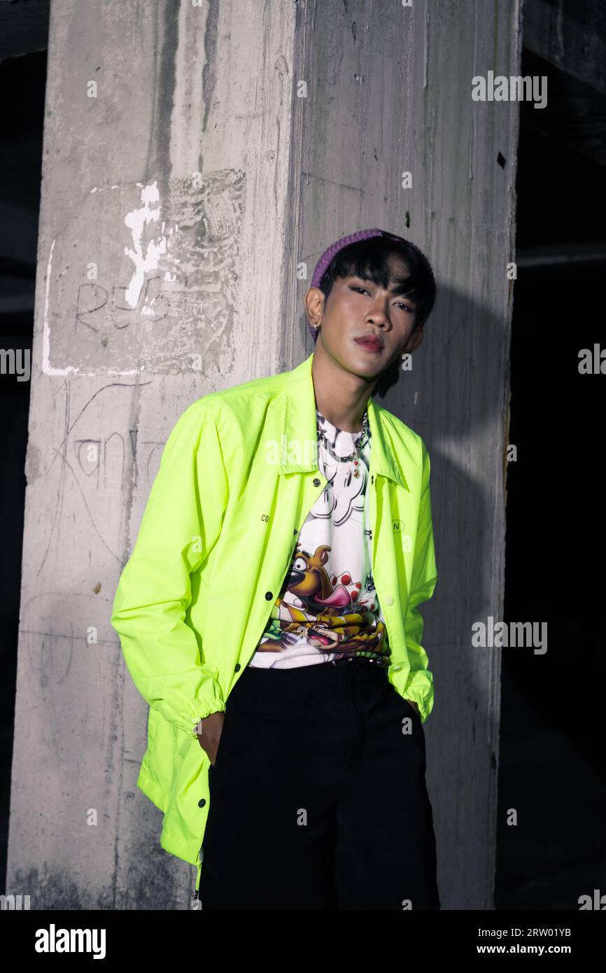 Ein asiatischer Mann in einer kalkfarbenen Jacke, die sich an einer stillgelegten Gebäudesäule lehnt und nachts sehr hübsch aussieht Stockfoto