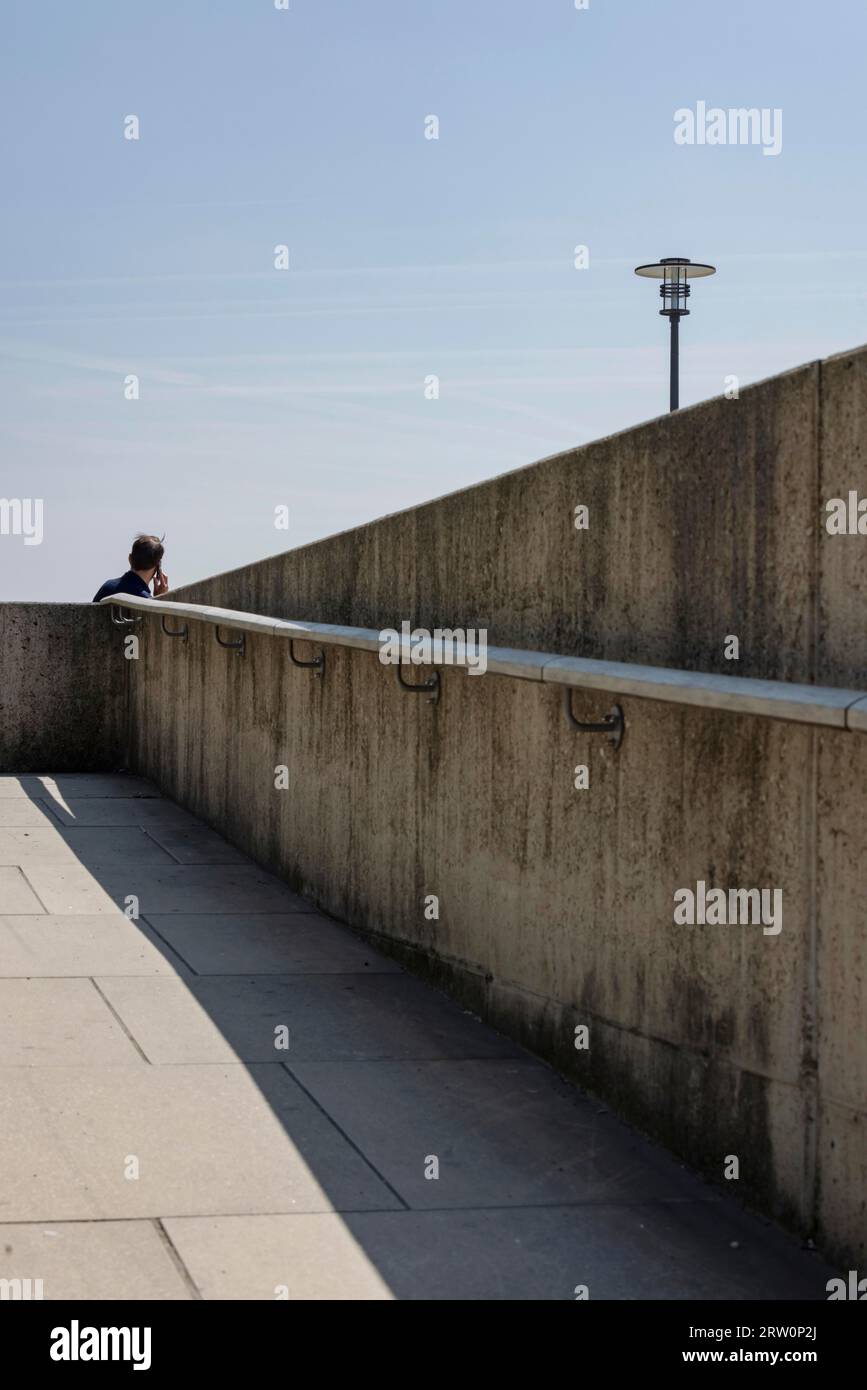 Mann, der auf seinem Smartphone auf einem sterilen Betonsteg zur Rheinpromenade in Köln, Nordrhein-Westfalen, Deutschland spricht Stockfoto