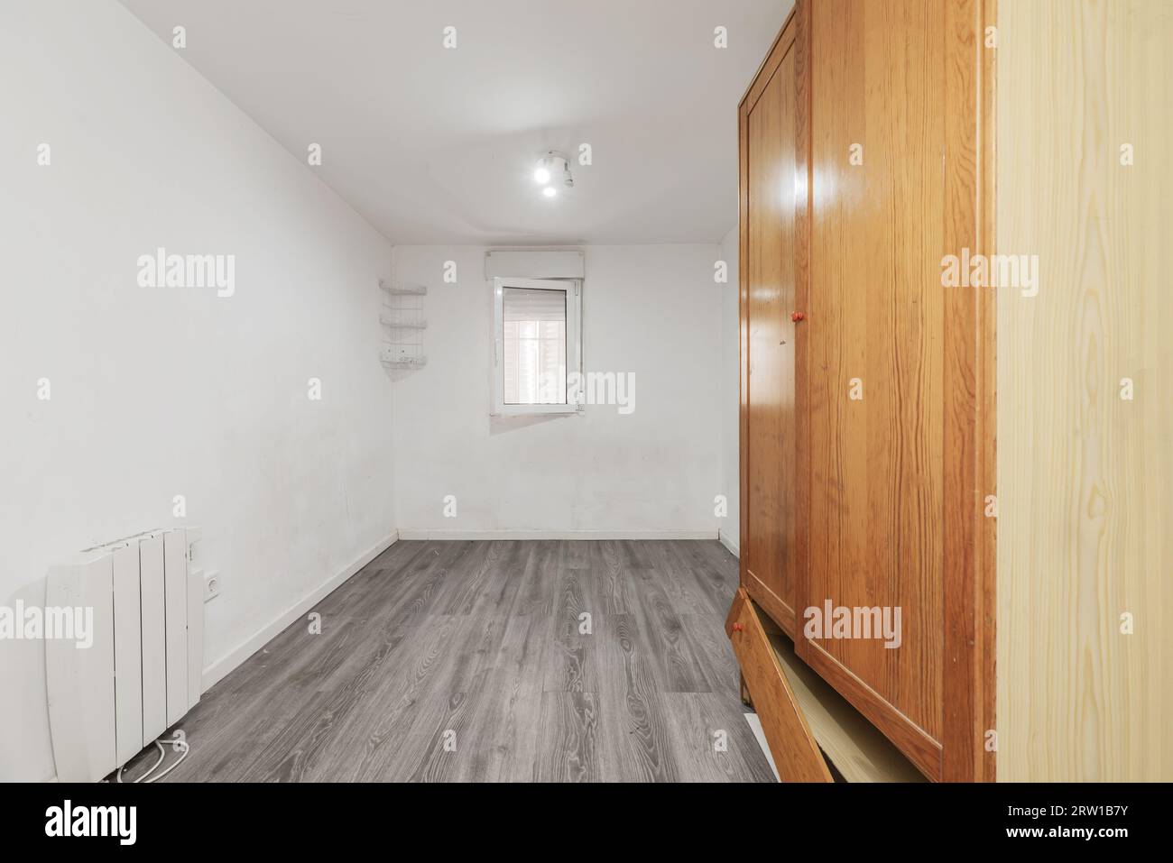 Leeres Zimmer mit einem wackeligen Kleiderschrank, elektrischem Heizkörper, grauem Holzfußboden, weißem Aluminiumfenster und weißen Wänden Stockfoto