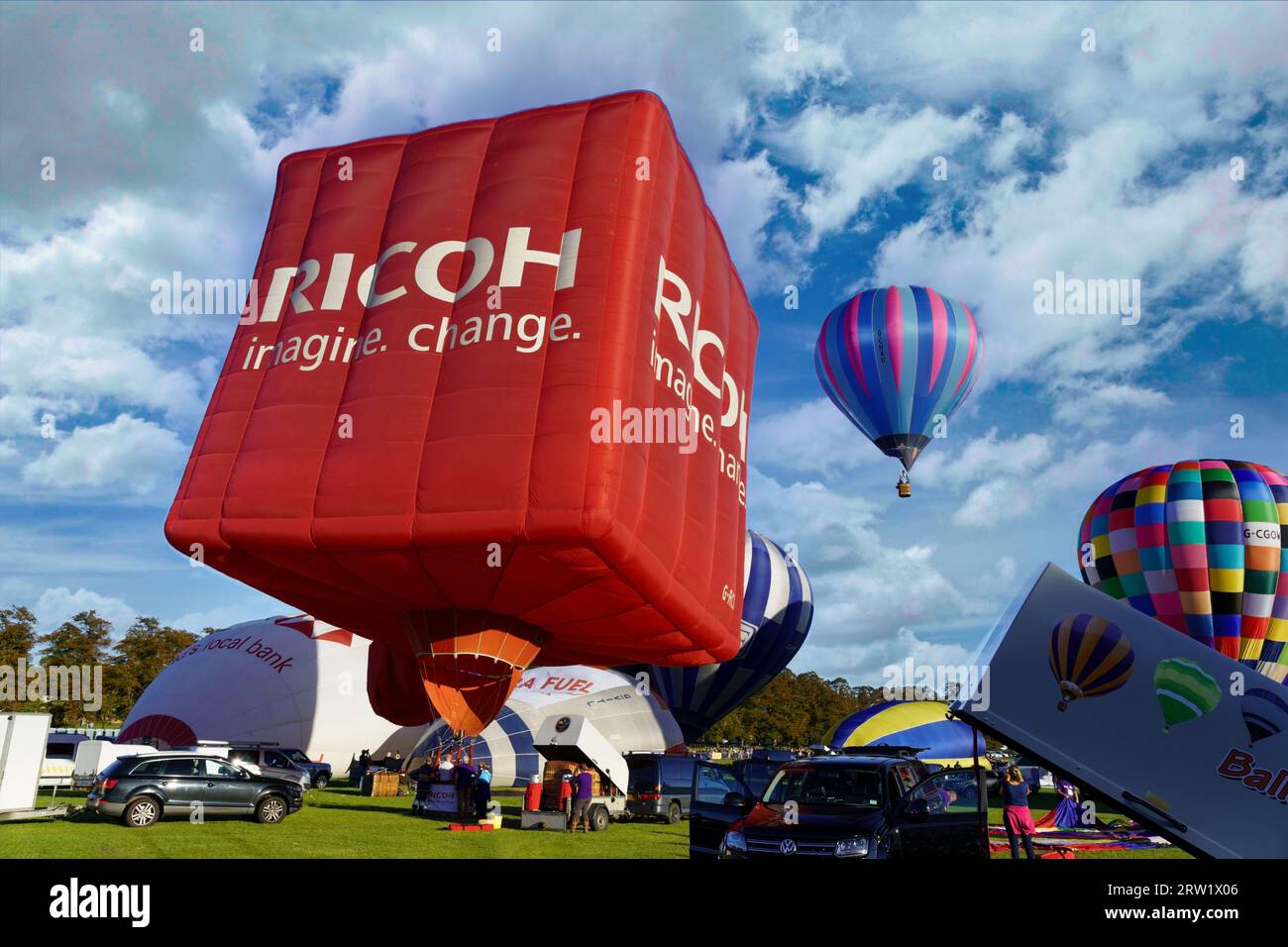 Würfelförmiger großer roter Ricoh-Heißluftballon, der sich auf den Start mit einem Ballon vorbereitet, der in der Ferne fliegt, York Balloon Fiesta, North Yorkshire, UK. Stockfoto