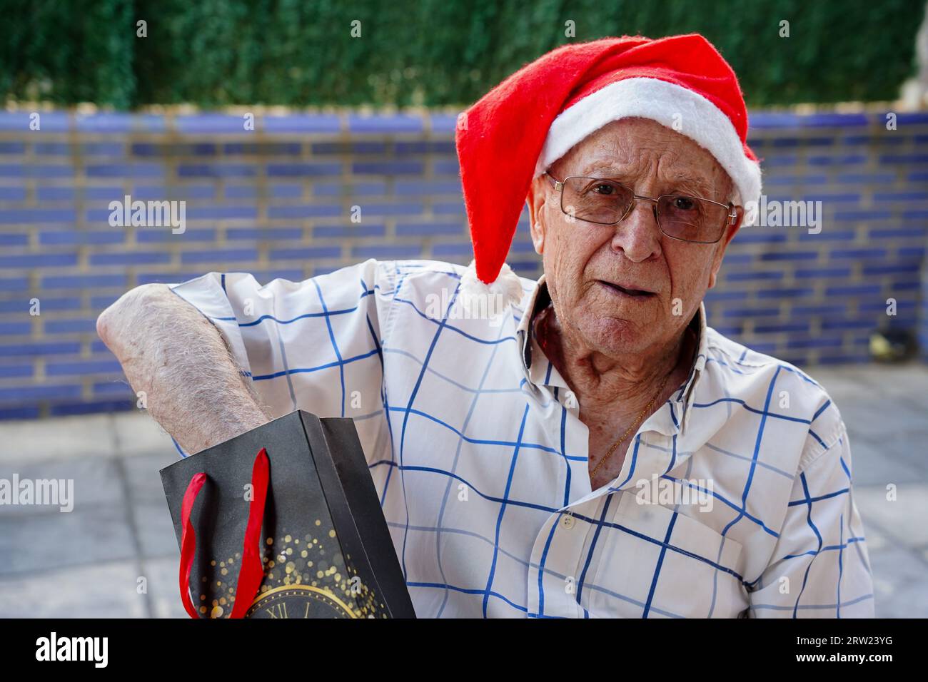 Älterer Mann mit Weihnachtsmann-Hut im Sommer. Der kaukasische Großvater öffnet ein weihnachtsgeschenk mit Blick auf die Kamera Stockfoto