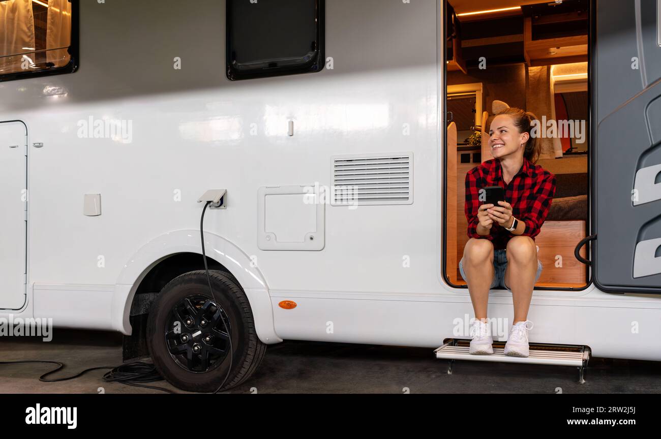 Glückliche junge Frau, die im Wohnmobil sitzt, benutzt ihr Handy, während der Van im Wohnmobil-Park auflädt. Haus auf Rädern. Vermietung von Wohnmobilen. Stockfoto