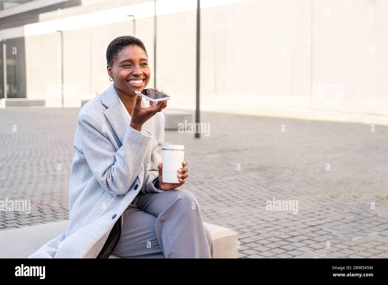 Horizontales Porträt einer eleganten jungen Afro-amerikanerin. Sie sitzt in der Nähe des Bürogebäudes und hinterlässt eine Sprachnachricht mit dem Telefon Stockfoto