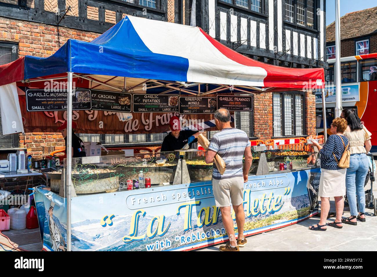 Leute am Lebensmittelmarkt kaufen französisches Tartiflette und andere Lebensmittel. Überdachung der französischen Flagge über dem Stand, im Hintergrund historisches Rathaus von Sandwich. Stockfoto