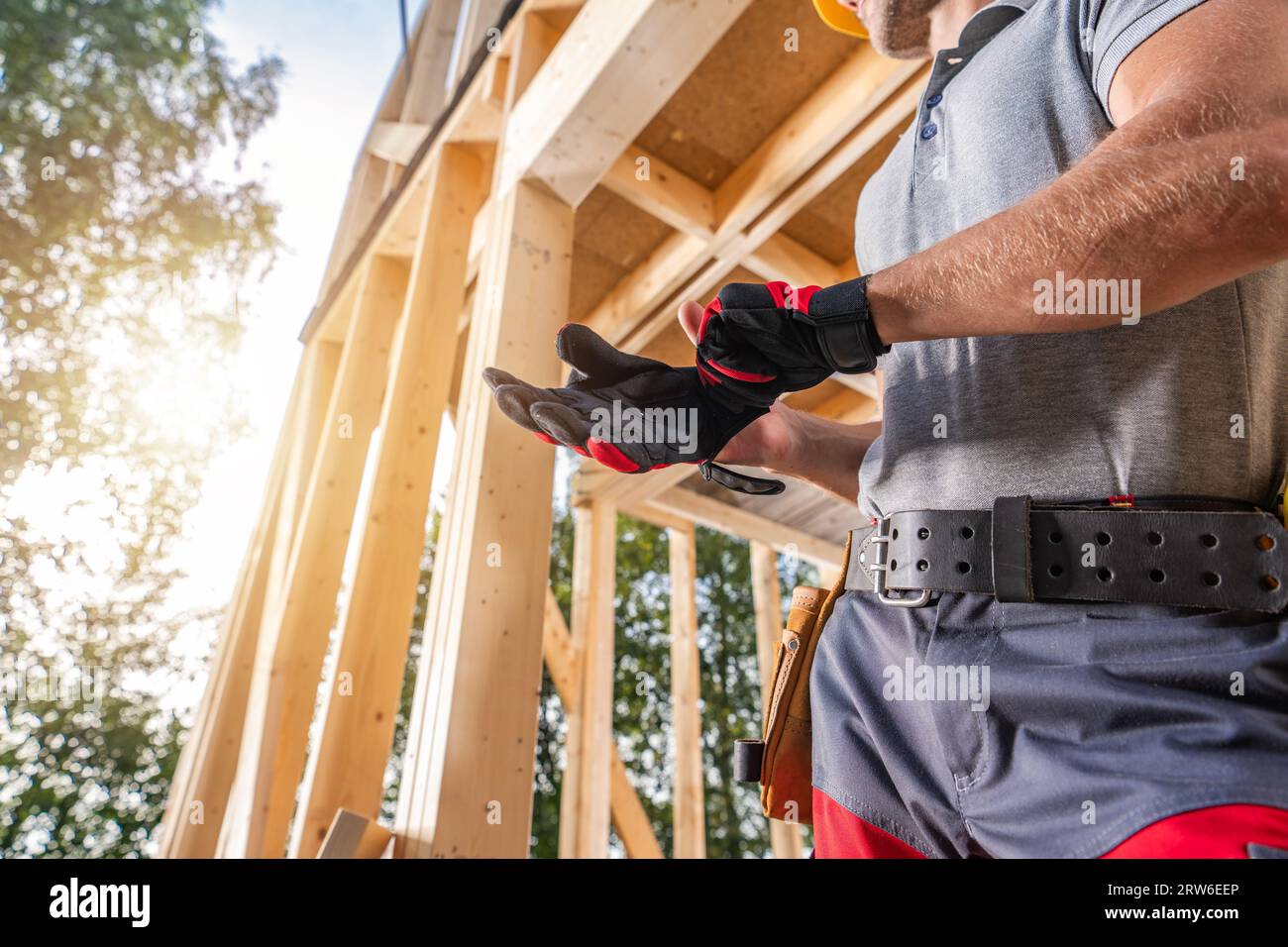 Bauunternehmer, der Schutzhandschuhe trägt, während er neben dem Holzhaus-Skelett-Rahmen bleibt Stockfoto