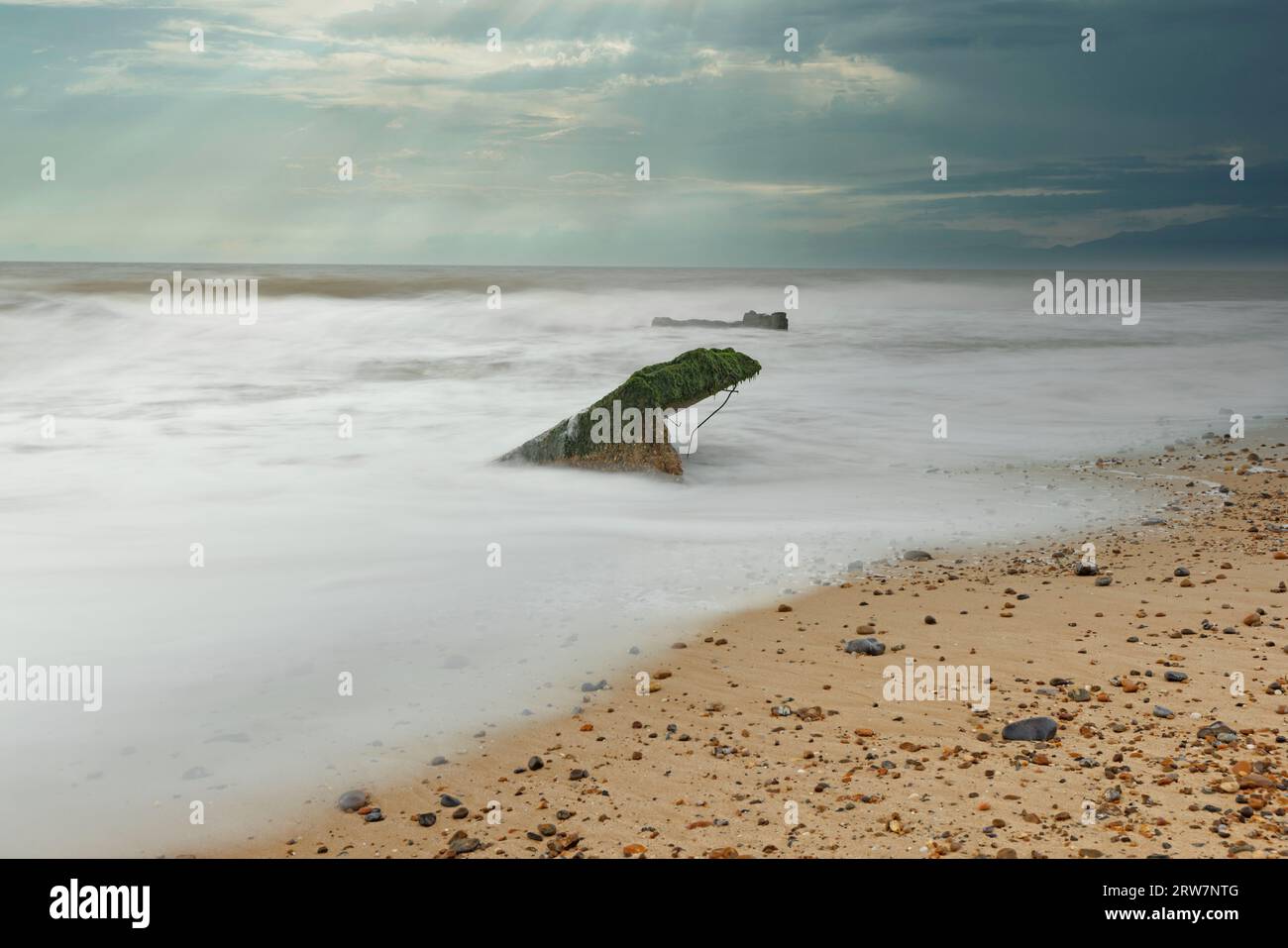 Ein Stück Treibholz im Sand, während die Wellen um ihn herum kreisen, und ein schöner bläulich-grüner Himmel dahinter Stockfoto