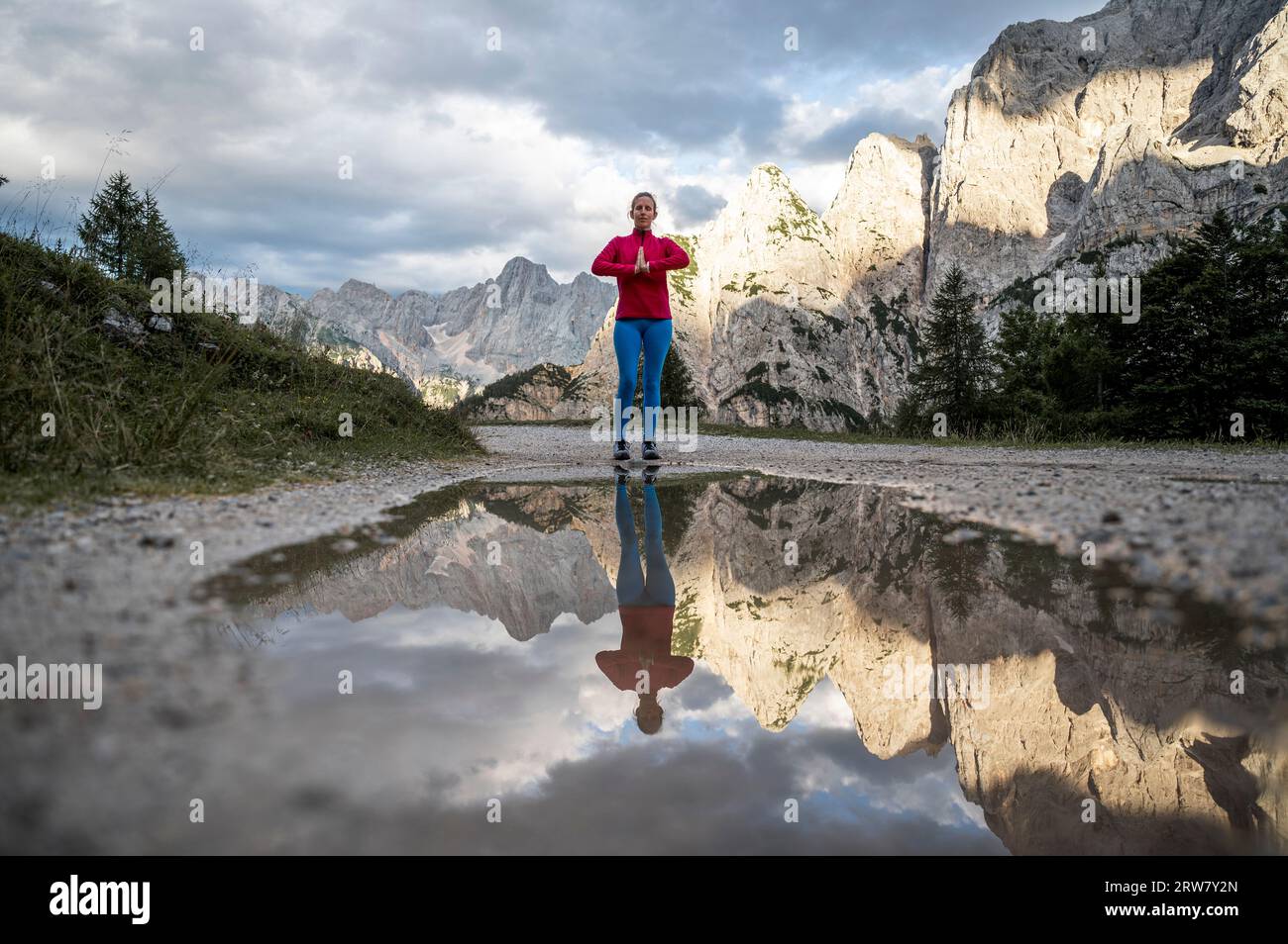 Junge Frau, die mit ihren Palmen meditierte, schloss sich vor ihrer Brust, während sie auf einem Weg stand, der von wunderschönen Bergen umgeben war. Mit einer Reflexion über ihn Stockfoto