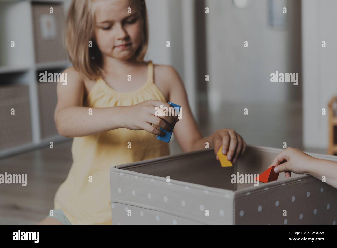 Kinder putzen Spielzeuge nach dem Spielen vom Boden und legen sie in eine Stoffkiste. Kinder sitzen auf Teppich im sonnigen weißen Schlafzimmer, Kindergartenspielzimmer oder p Stockfoto