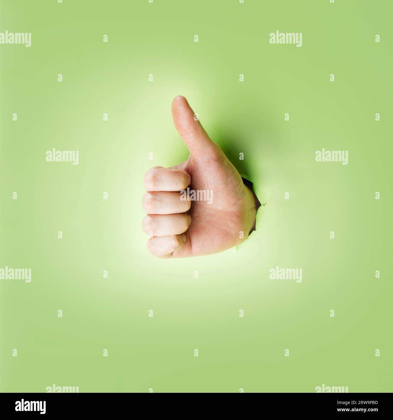 Hand macht eine Daumen-hoch-Geste durch ein Loch in grünem Papier Stockfoto