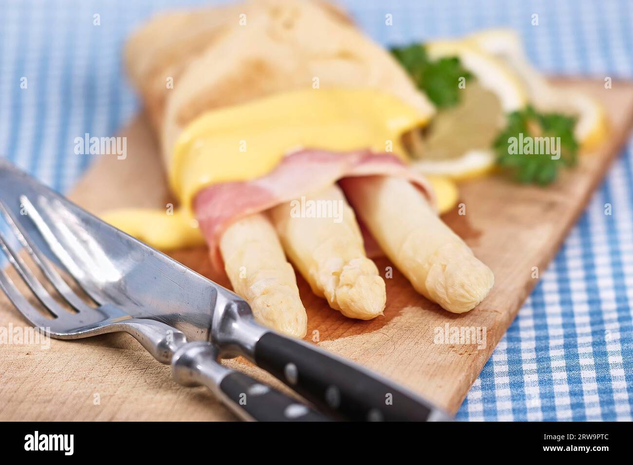 Weißer Spargel mit Schinken und Omelette liegt auf einem rustikalen Holzbrett. Rustikales Besteck liegt links vorne. Das Bild hat ein feines Bokeh Stockfoto