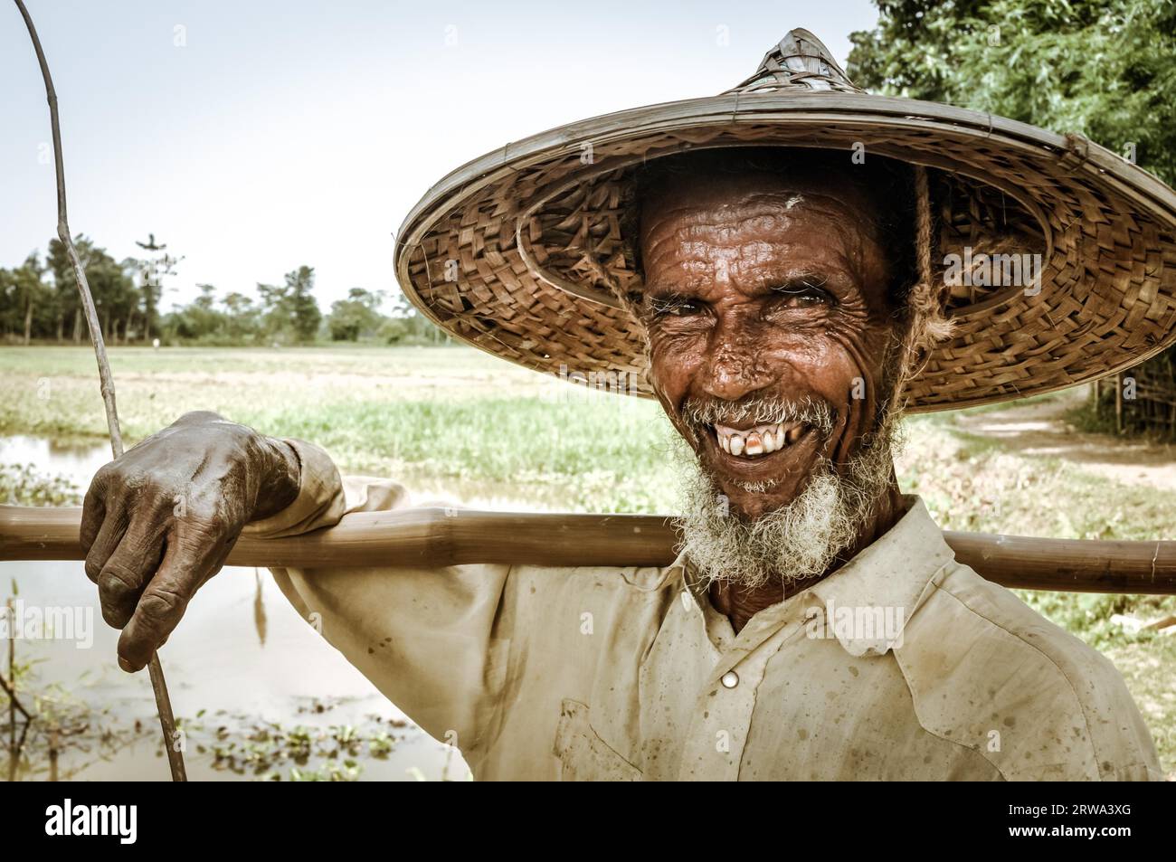 Srimongal, Bangladesch, um Juli 2012: Der alte Mann mit grauem Bart und großem Strohhut hält in Srimongal, Bangladesch, einen Holzstock auf der Schulter. Stockfoto