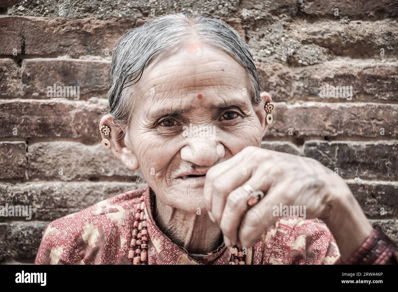 Bhaktapur, Nepal, um Juni 2012: Alte grauhaarige Frau mit faltigem Gesicht in roten Kleidern mit Kette aus Perlen trägt Ohrringe in Stockfoto