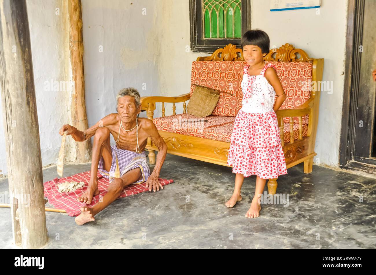 Srimongal, Bangladesch, um Juli 2012: Der alte dünne Mann sitzt auf dem Boden und seine Enkelin in rot-weißem Kleid steht in Srimongal in seiner Nähe Stockfoto