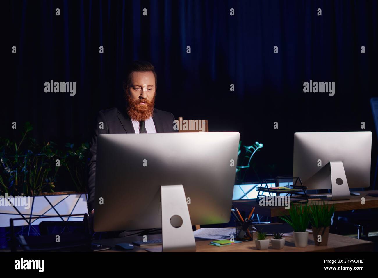 Seriöser bärtiger Geschäftsmann, der sich den Computermonitor anschaut, während er spät im Büro arbeitet, Nachtschicht Stockfoto