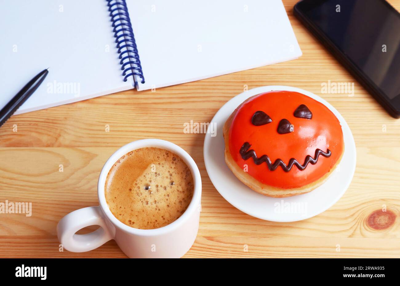 Tasse heißer Kaffee mit einem leuchtend orangefarbenen Halloween-Donut auf einem Schreibtisch Stockfoto