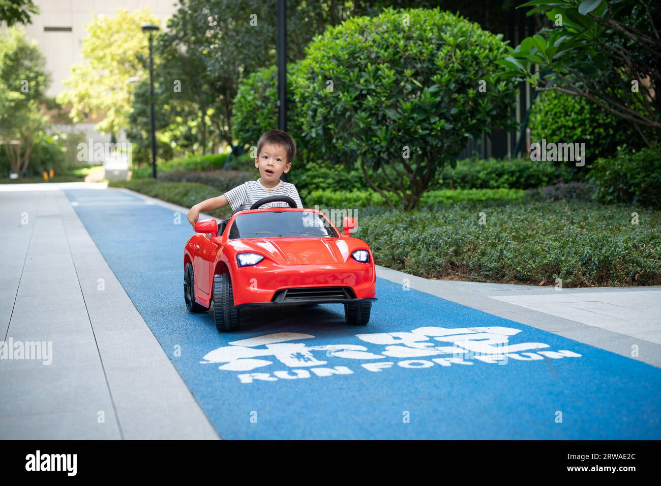 Der süße, multirassische Kleinkind schaut sich um, während er mit einem roten ferngesteuerten Auto auf einer befestigten Strecke fährt Stockfoto