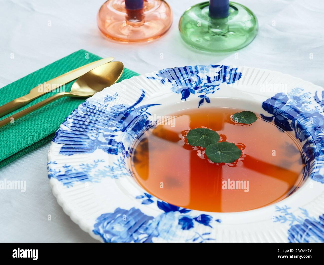 Nahaufnahme eines sonnenverwöhnten Mittagesstisches mit einer porzellanschale im Delfter Stil mit dünner Tomatenconsommé-Suppe oder leichter Brühe, garniert mit Kapuzinerkressblättern Stockfoto