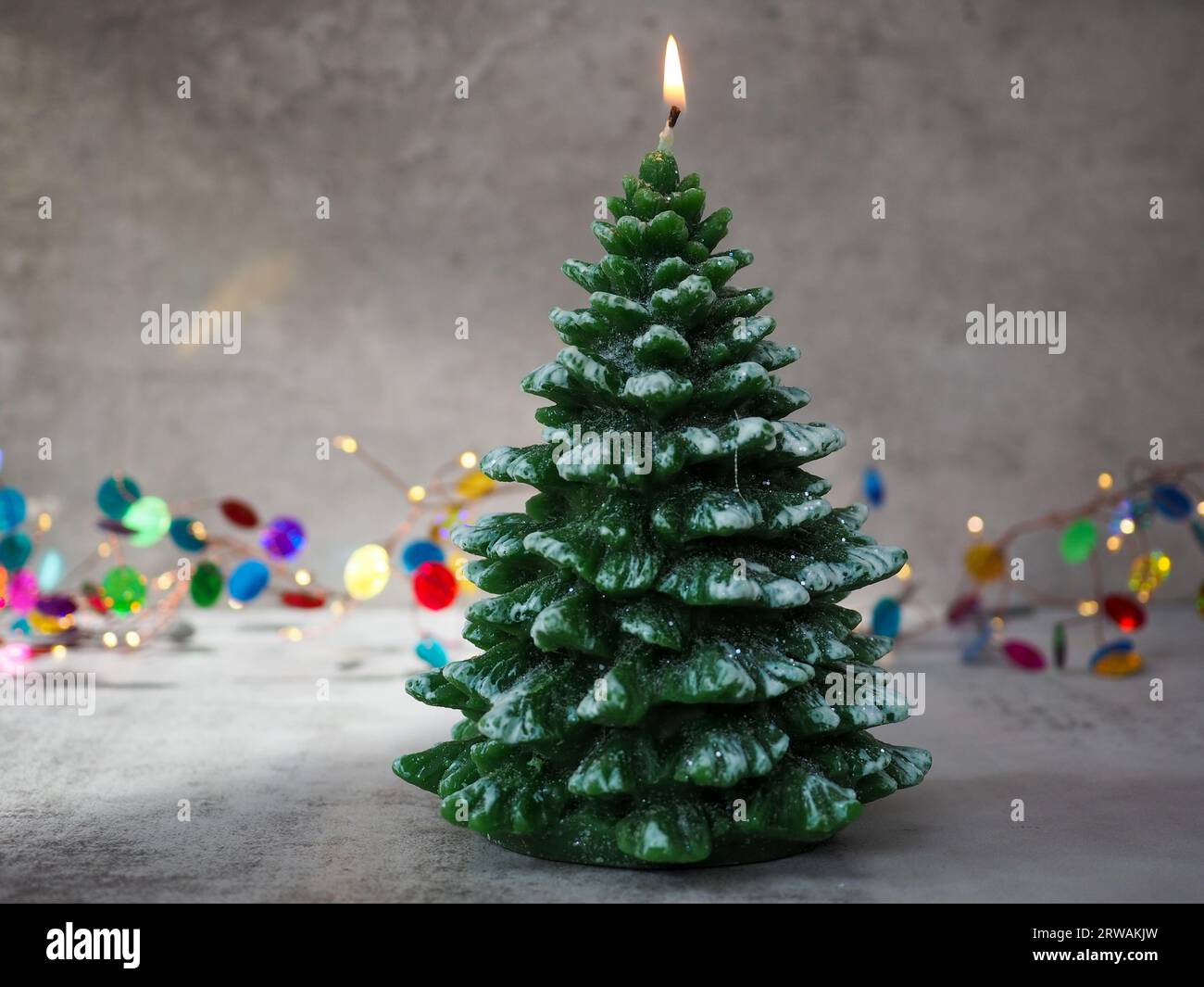 Eine beleuchtete Kerze in Form eines Weihnachtsbaums vor dem Bokeh einer farbenfrohen Lichterdekoration mit einem betonierten grauen Hintergrund und Platz für Kopien Stockfoto
