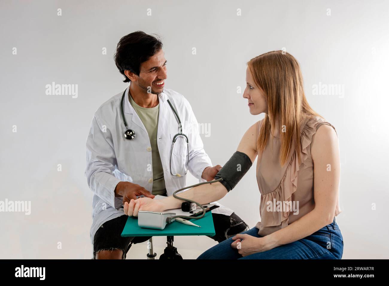 Asiatischer Arzt aus Indien, gekleidet in einem weißen Mantel, der den Blutdruck an einer rothaarigen europäischen Frau misst. Stockfoto