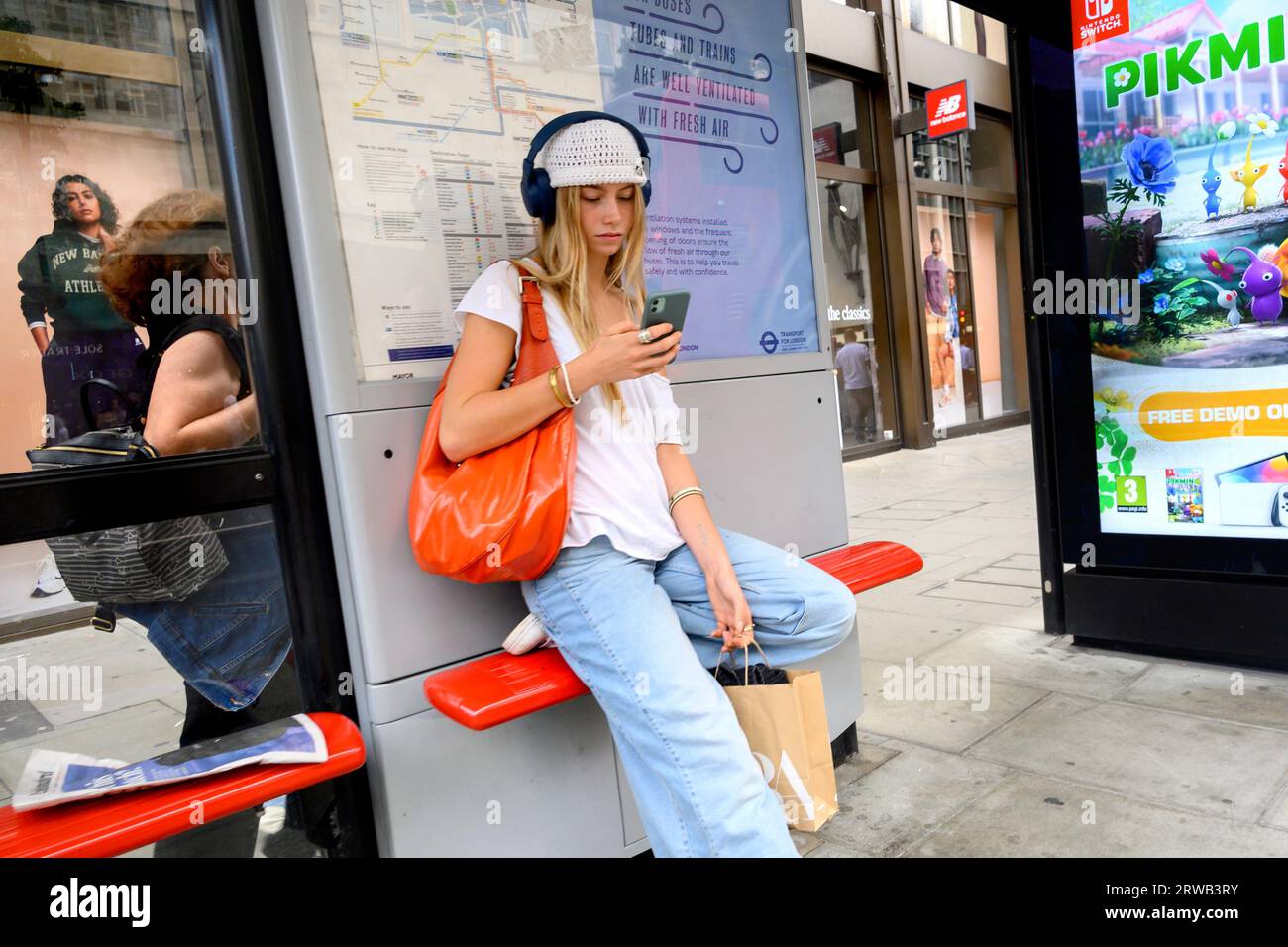 London, Großbritannien. Eine junge Frau an einer Bushaltestelle, die Kopfhörer trägt und ihr Handy ansieht Stockfoto