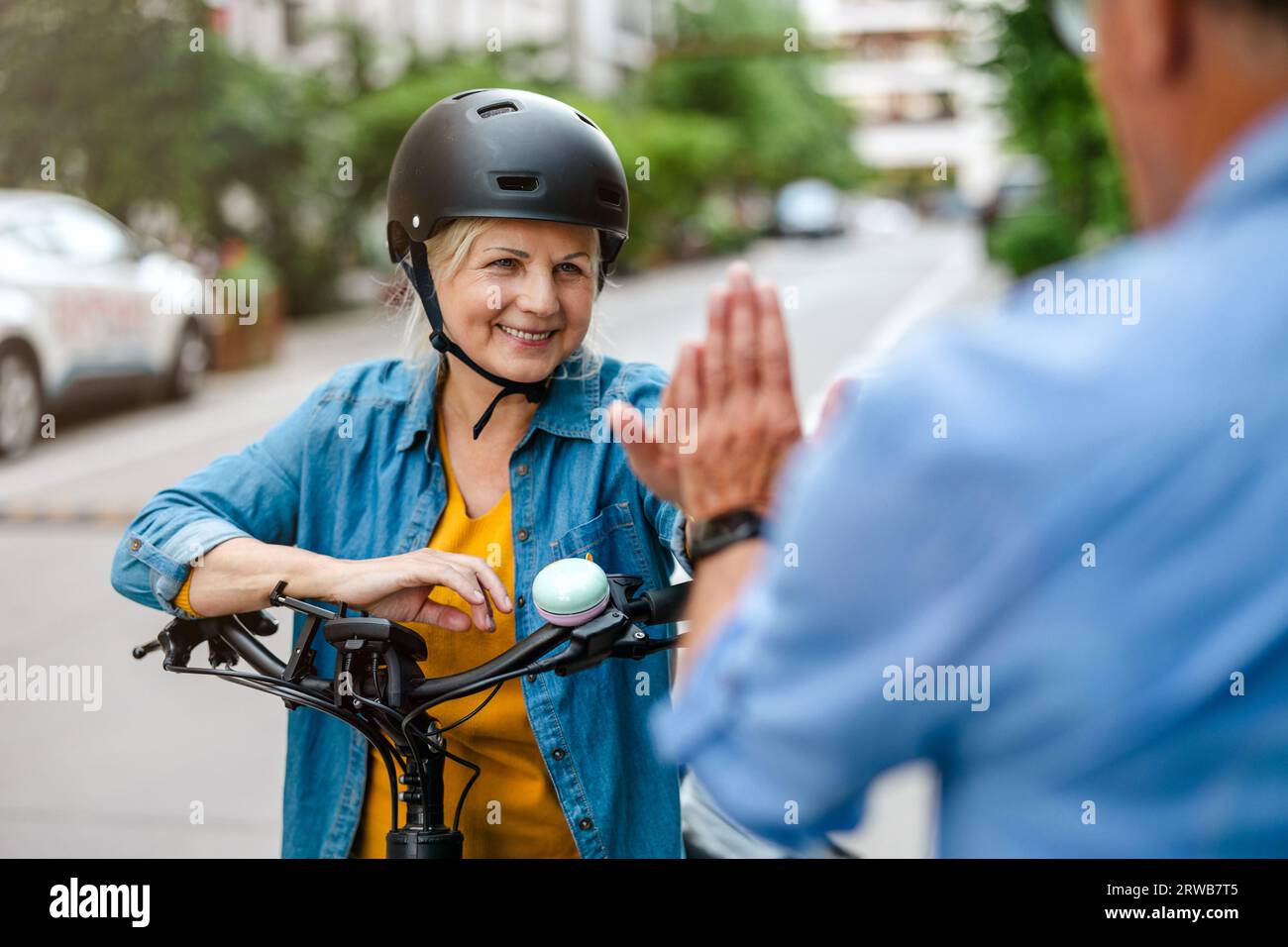 Lächelnde, reife Frau im Helm, die dem Mann beim Fahrradfahren hohe fünf schenkt Stockfoto