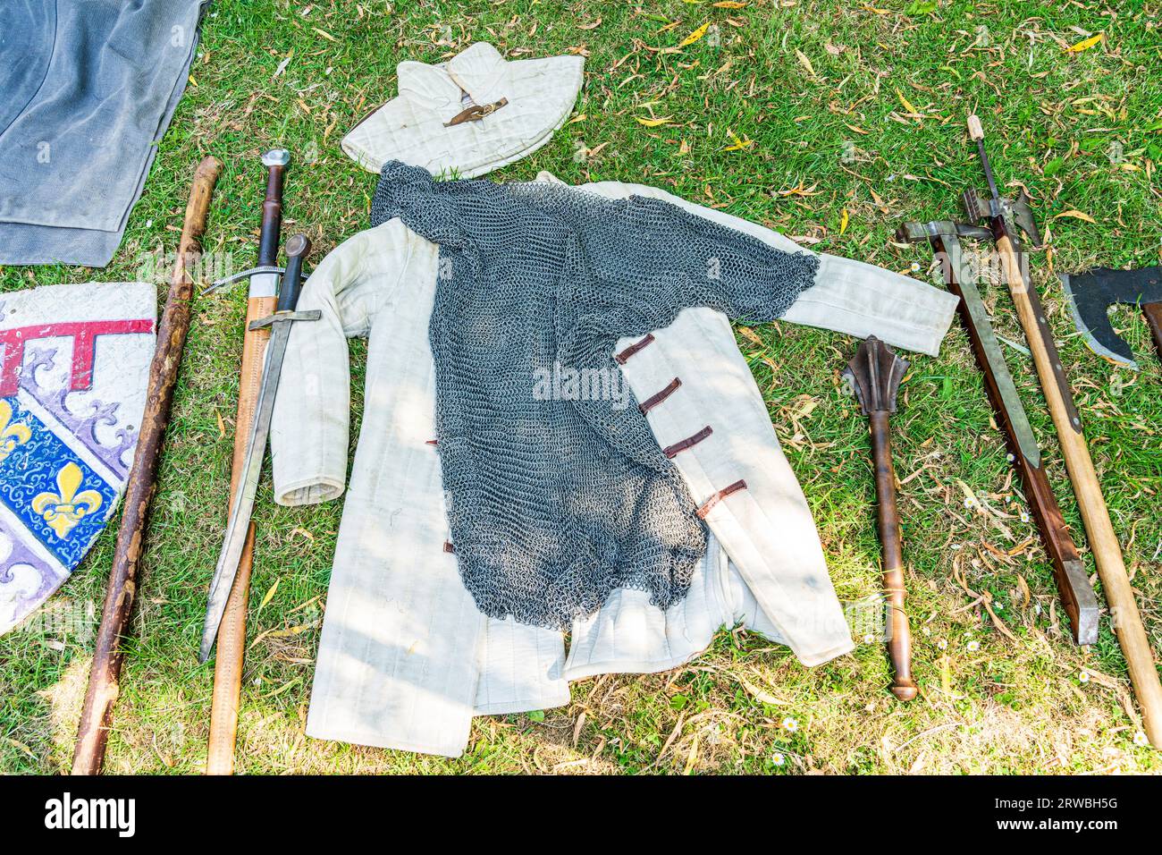 Mehrere mittelalterliche Waffen, Schwerter, Streitkolben und Kampfaxt, die um einen Anzug aus Kettenpost und Gambeson angeordnet waren, ein unter dem Gras getragenes Kleid. Stockfoto