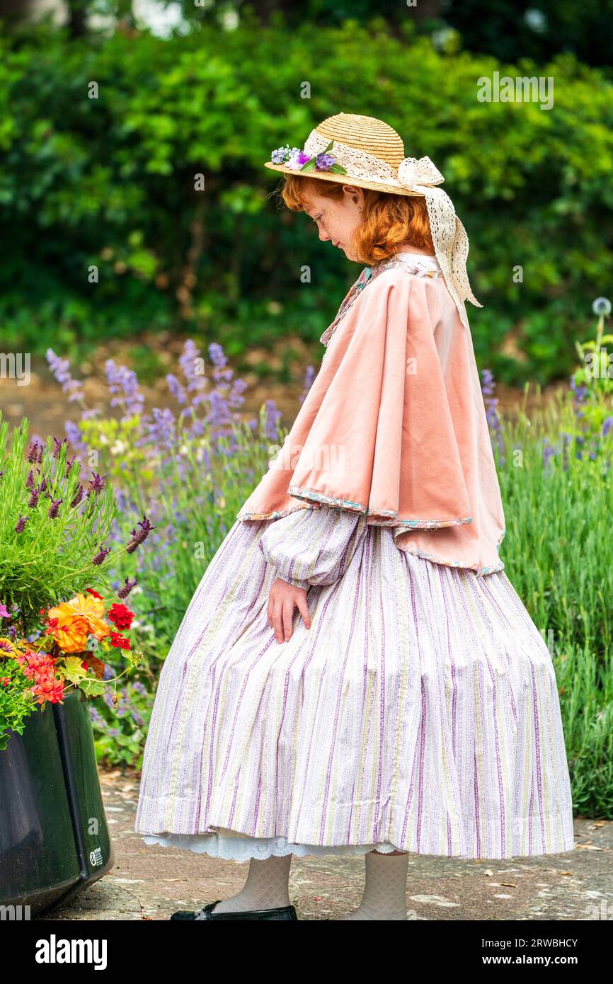 Junge Teenager-Frau im Victoria-Kostüm, die traurig auf einige Blumen in einer Plastikschale in einem Garten schaut. Sie trägt einen Strohhut, ein Tuch und einen weiten Rock. Stockfoto